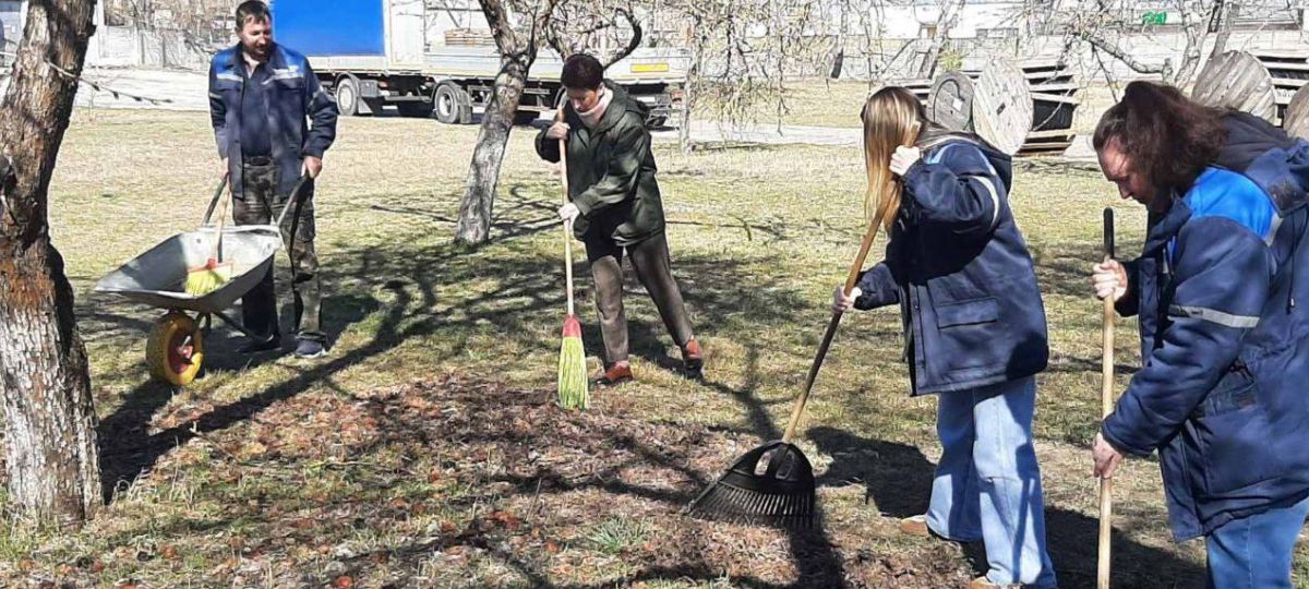 Жители в рабочей одежде убирают территорию: сгребают листву граблями и метлой, рядом стоит тачка
